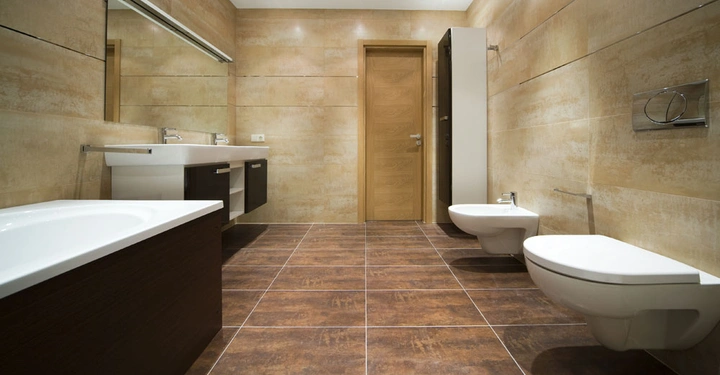 Badezimmer mit braunen Fliesen an Boden und Wänden Doppelwaschbecken links Badewanne mit Holzverkleidung Dusche rechts sowie Toilette und Bidet Tür in der Mitte beleuchtet durch Deckenlichter