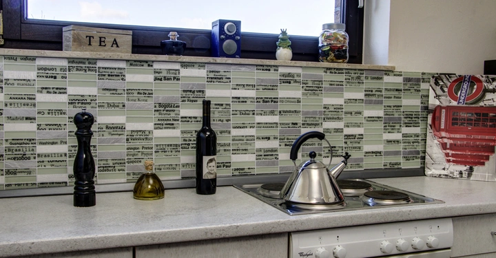 Ein Edelstahl-Wasserkocher steht auf einem Herd in einer modernen Küche neben einer Weinflasche Pfeffermühle und Ölbehälter. Über dem Herd ist ein Fliesenspiegel mit Städte- und Zeitungsnamen.