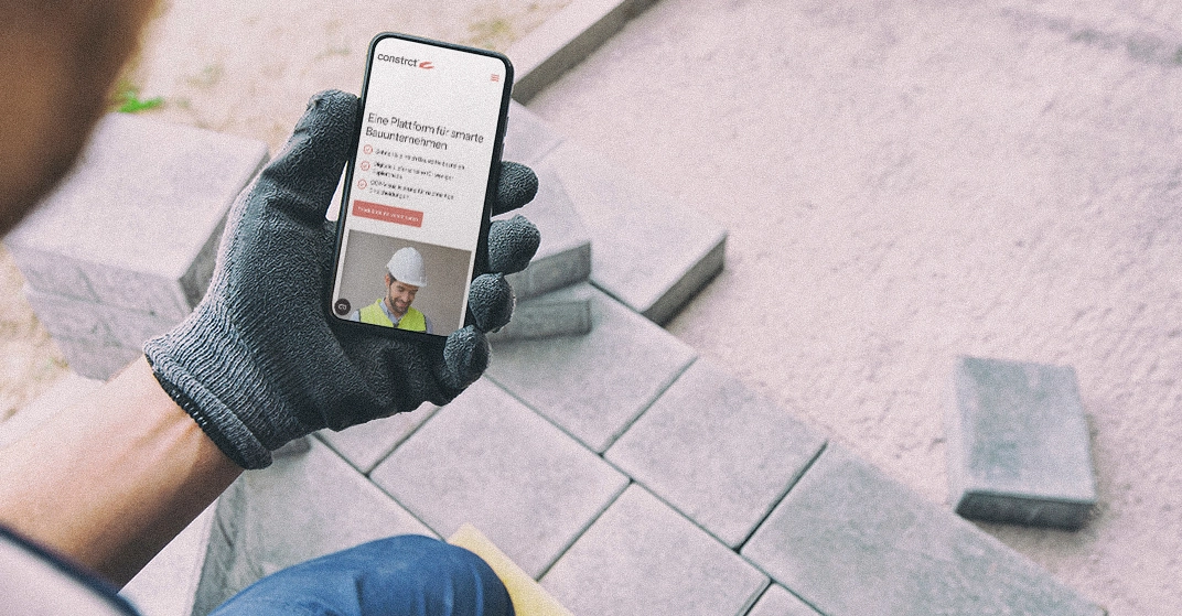 Mann mit Arbeitshandschuhen schaut auf Handy und hat dort die App constrct geöffnet.
