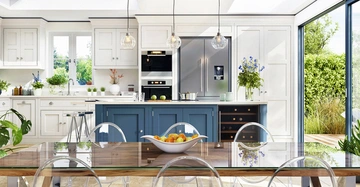 Offene Küche im Landhausstil mit weißen und blauen Küchenkästen in Frontalansicht. Rechts Ausgang auf Terrasse. Monstera links am Boden, zahlreiche Kräuter und Zimmerpflanzen auf Regalen.