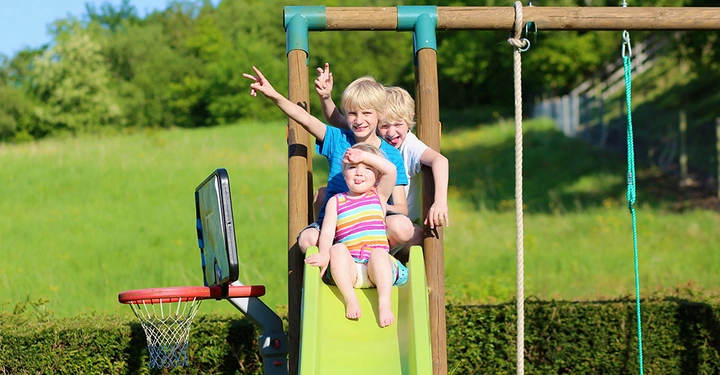 Spielturm aus Holz mit hellgrüner Rutsche, oben sitzen drei kleine Kinder hintereinander, Arme fröhlich in die Luft gestreckt. Hochsommerlich, saftiger grüner Rasen, Bäume, blauer Himmel, Sonne