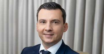 Mann lächelt trägt einen blauen Anzug und ein weißes Hemd steht vor einem Hintergrund aus hellen Vorhängen