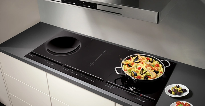 Ein schwarzes Induktionskochfeld mit einem Kochtopf voll bunter Paella auf der rechten Seite und zwei Tellern mit frischem Gemüse daneben in einer modernen Küche.