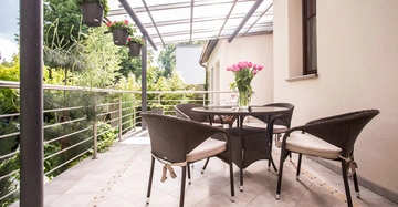 Ein runder Tisch mit vier Stühlen und einem Blumenstrauß sitzt auf einer überdachten Terrasse mit Blick auf einen grünen Garten. Hängende Pflanzen schmücken die Geländer.