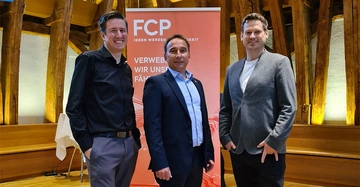 Drei Männer lächeln und posieren vor einem orangefarbenen Banner mit der Aufschrift FCP Ideen werden Wirklichkeit Verweben wir unsere Fähigkeiten in einem hölzernen Raum mit Bänken und Leuchten im Hintergrund
