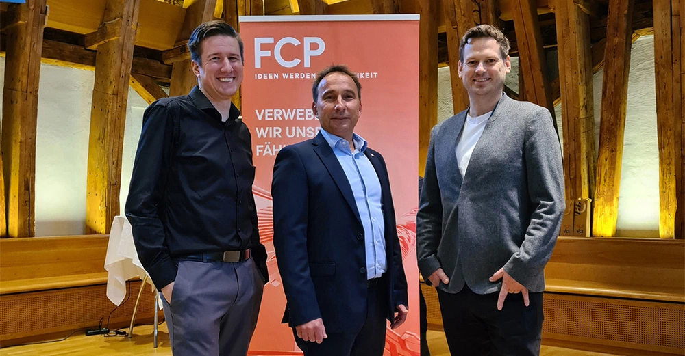 Drei Männer lächeln und posieren vor einem orangefarbenen Banner mit der Aufschrift FCP Ideen werden Wirklichkeit Verweben wir unsere Fähigkeiten in einem hölzernen Raum mit Bänken und Leuchten im Hintergrund