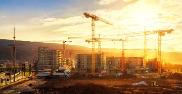 Krane heben Baumaterialien auf einer Baustelle mit halbfertigen Gebäuden umgeben von Bergen unter einem Sonnenuntergangshimmel