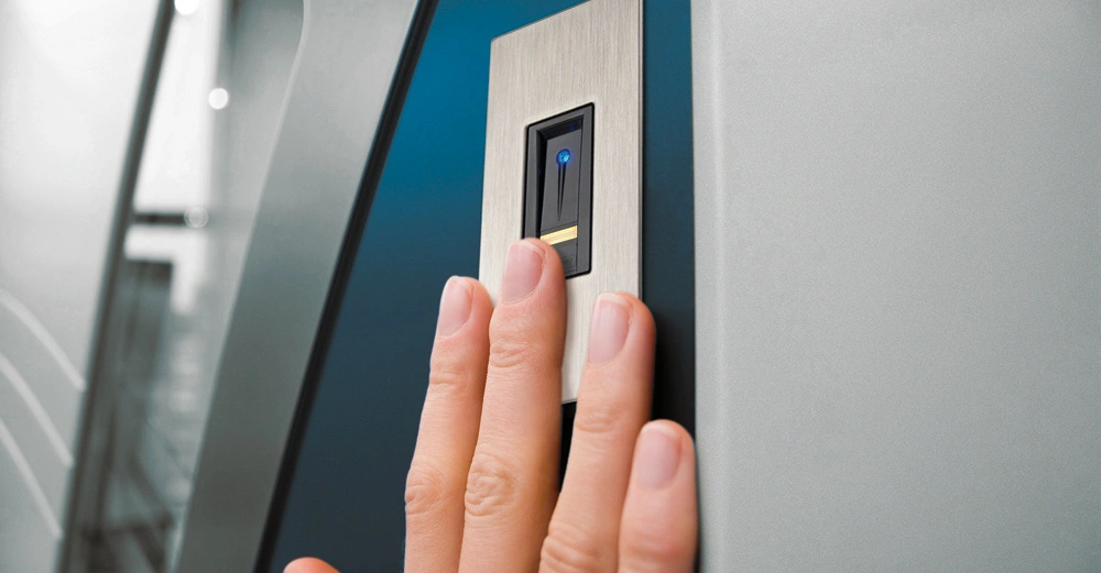 Hand drückt auf Fingerabdruckscanner an einer metallischen Oberfläche in einem modernen, beleuchteten Raum.