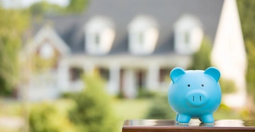 Blaues Sparschwein steht auf einem Tisch. Im Hintergrund unscharf ein großes Haus mit weißer Fassade und mehreren Fenstern. Grüne Bäume und Sträucher umgeben das Haus.