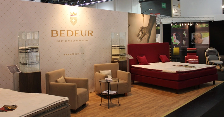 Galerie-Bild 2: Zwei Betten und zwei Sessel stehen auf einem Holzboden vor einer Ausstellungswand mit dem Schriftzug BEDEUR FIRST CLASS LUXURY SLEEP WWW BEDEUR COM in einer beleuchteten Messehalle.