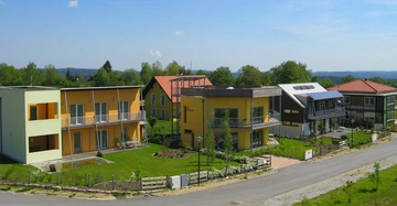 Mehrere moderne Einfamilienhäuser stehen nebeneinander in einer ruhigen Wohnsiedlung mit gepflegten Gärten und einer asphaltierten Straße bei sonnigem Wetter im Grünen.
