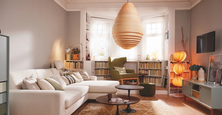 Ein beige Sofa steht in einem Wohnzimmer eingerahmt von Bücherregalen. Eine große hölzerne Hängelampe hängt von der Decke. Der Raum ist hell und gemütlich eingerichtet.