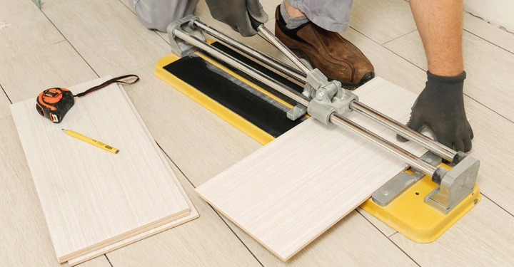 Eine Person benutzt einen Fliesenschneider um eine Fliese zu schneiden auf einem gefliesten Boden daneben ein Maßband und ein Bleistift auf anderen Fliesen.