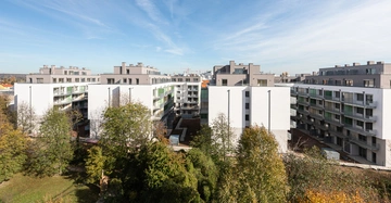 Mehrere moderne Wohngebäude stehen nebeneinander. Im Vordergrund befindet sich ein grüner Park mit Bäumen. Der Himmel ist blau mit einigen Wolkenstreifen.