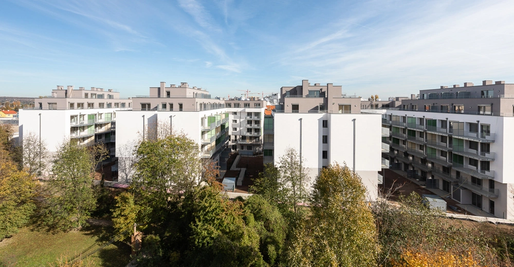 Mehrere moderne Wohngebäude stehen nebeneinander. Im Vordergrund befindet sich ein grüner Park mit Bäumen. Der Himmel ist blau mit einigen Wolkenstreifen.