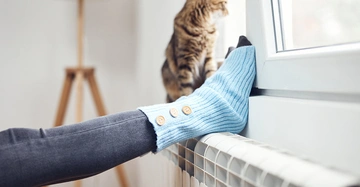 Frau lehnt mit dem Füßen auf einem Heizkörper unter einem modernen Fenster. Eine braune Tigerkatze sitzt auf der Fensterbank und sieht nach draußen.
