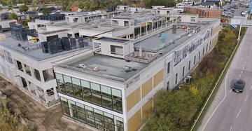 Ein großes Gebäude im Bau sammelt Baumaterialien auf seinem Dach und ist von einer Straße mit einem vorbeifahrenden Auto umgeben. Mehrere kleinere Gebäude sind im Hintergrund sichtbar.