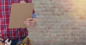 Person hält ein Klemmbrett und trägt ein rot-kariertes Hemd im Vordergrund Baumarkt-Werkzeuge am Gürtel Hintergrund eine unscharfe Ziegelsteinwand.
