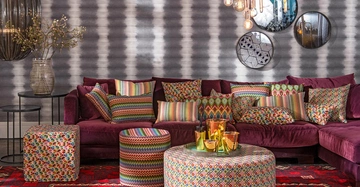 L-förmiges, violettes Sofa mit bunten Kissen und Hockern steht in modernem Wohnzimmer mit grauer, gestreifter Tapete Runder, gemusterter Couchtisch und rote Teppiche vervollständigen den Raum. Hängende Glühbirnen und drei runde Spiegel an der Wand.