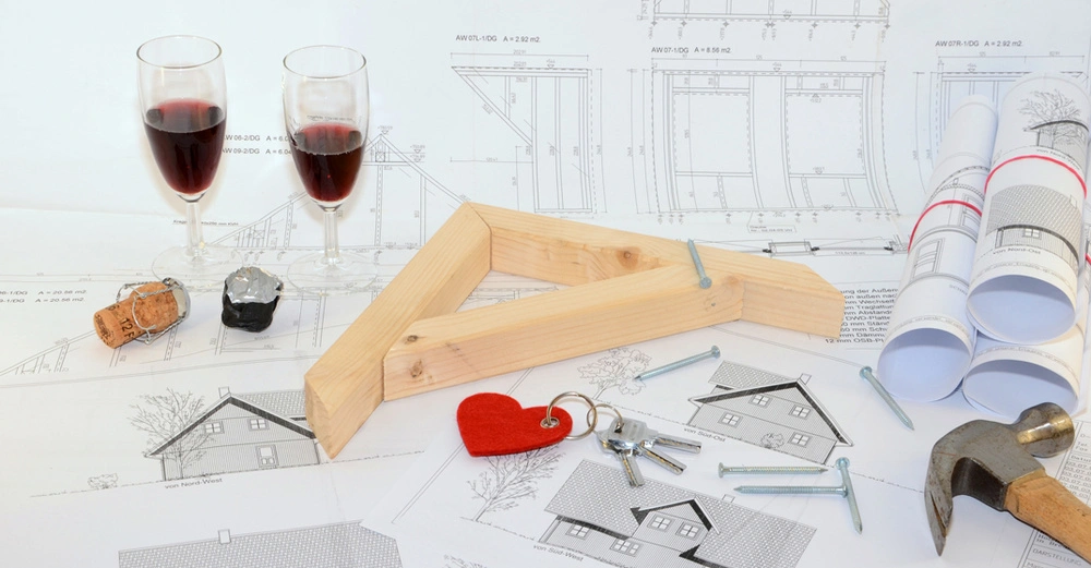 Hauspläne liegen auf einem Tisch, darauf ein Modell eines Hauses aus Holz, Messstab, zwei Sektgläser und ein Haustürschlüssel. 
