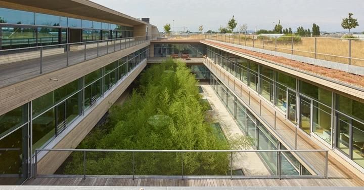 Galerie-Bild 5: Mehrstöckiges modernes Gebäude mit vielen Glasfenstern, grüne Pflanzen im Innenhof, Holzverkleidung und Kies auf dem Dach, umgeben von Bäumen am Horizont unter leicht bewölktem Himmel.