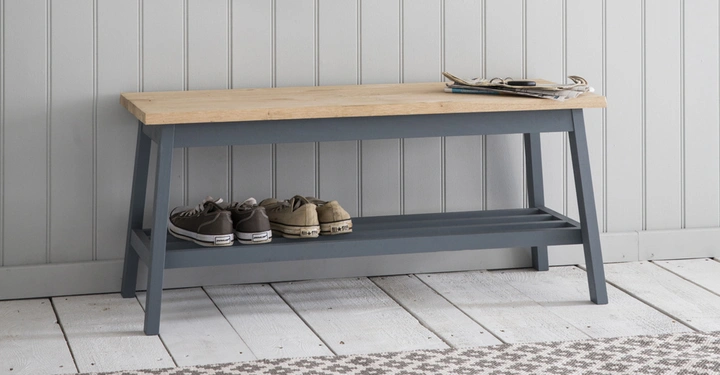 Holzbank steht still, darauf liegen Zeitschriften, darunter befinden sich Schuhe; die Bank steht in einem Raum mit weißer, vertäfelter Wand und weißem Holzboden, auf dem ein Teppich liegt.