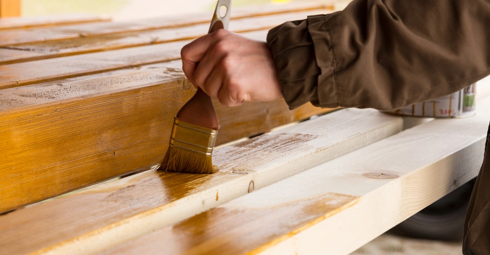 männliche Hand pinselt nebeneinanderliegende Holzbalken mit Holzschutzmittel ein 