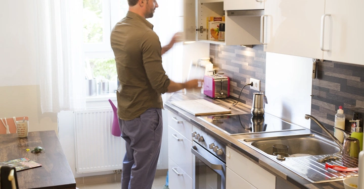Galerie-Bild 4: Ein Mann öffnet einen Küchenschrank und nimmt etwas heraus in einer gut beleuchteten Küche mit einem Esstisch und einem großen Fenster im Hintergrund.