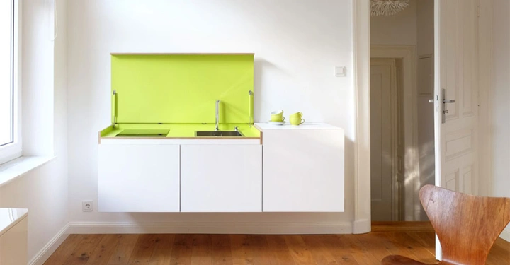 Weiße Küchenschränke mit grünem Rückwand-Paneel und Spüle eingebaut an weißer Wand in einem Raum mit Holzboden und Holzstuhl am rechten Rand eines hellen Raumes mit Tür und Fenster