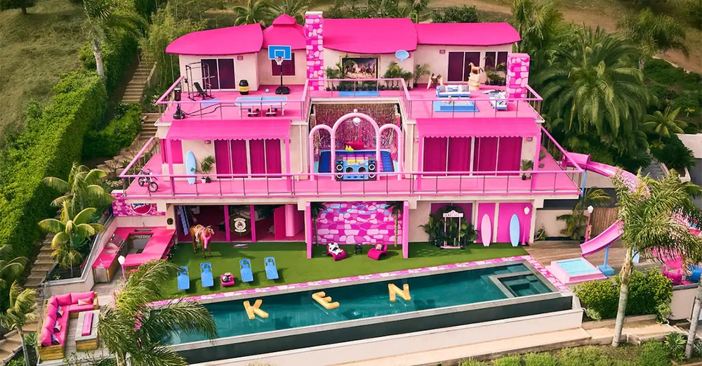 Großes mehrstöckiges Barbiehaus in Pink mit mehreren Balkonen und einer Dachterrasse umgeben von Palmen Schwimmbad im Vordergrund mit "KEN" in gelben Buchstaben auf dem Wasser