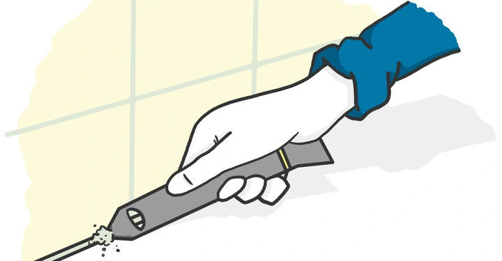 Eine Hand in einem blauen Ärmel hält ein Werkzeug und entfernt Fugenmaterial zwischen Fliesen an einer Wand.