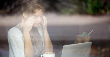 Eine Frau sitzt in einem Café vor einem Laptop und massiert sich die Schläfen. Vor ihr steht eine Tasse Kaffee. Durch das Fenster sind Bäume im Hintergrund sichtbar.