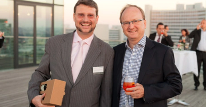 Galerie-Bild 34: Zwei Männer lächeln auf einer Dachterrasse Mit einer Hand hält der eine eine Box der andere ein Getränk Im Hintergrund sind andere Personen und Gebäude zu sehen.