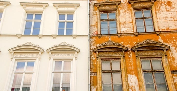 Zwei nebeneinander stehende Hausfassaden Eine ist frisch renoviert und hellweiß Die andere ist alt und abgenutzt mit gelb-bröckelnder Farbe Beide Fassaden haben identische Fensteranordnungen und Verzierungen