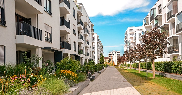 Moderne Wohnhäuser mit Balkonen stehen sich entlang eines gepflasterten Gehwegs gegenüber umgeben von grünen Pflanzen und Bäumen bei klarem Himmel im städtischen Wohngebiet.