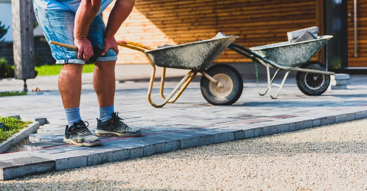 Mann kniet mit Werkzeug auf Pflastersteinen während er einen Gehweg legt zwei Schubkarren gefüllt mit Baumaterialien im Hintergrund sitzt vor einem Gebäude mit Holzverkleidung im Freien