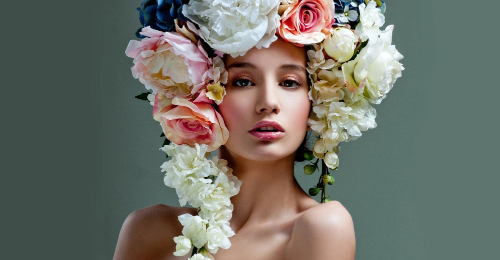 Eine Frau trägt einen auffälligen Kopfschmuck aus großen, bunten Blüten. Ihr Oberkörper ist unbekleidet und der Hintergrund ist grau.