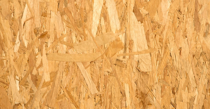 Holzpartikel sind zufällig verstreut auf einer Oberfläche. Keine Textur oder Muster; erscheint als grobe Holzwerkstoffplatte in Nahaufnahme. Die Umgebung ist neutral ohne erkennbare Zusatzgegenstände.