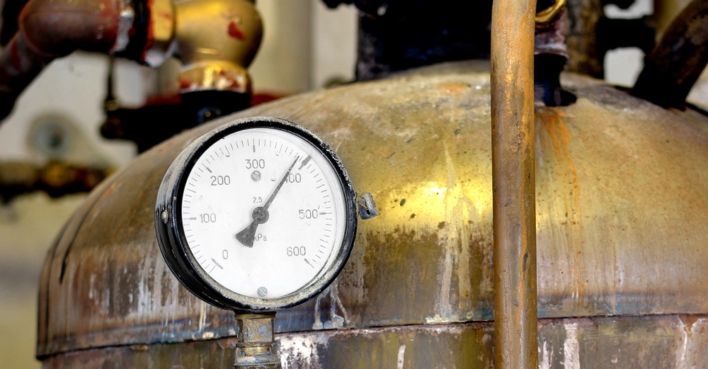 Ein Rundinstrument zeigt einen Druck von 2.5 MPa an und befindet sich oberhalb eines rostigen Metallbehälters in einer industriellen Umgebung mit verschiedenen Rohren und Armaturen im Hintergrund.