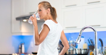 Frau trinkt Wasser aus einem Glas in der sauberen hellen Küche mit Küchengeräten und weißblauer Einrichtung.