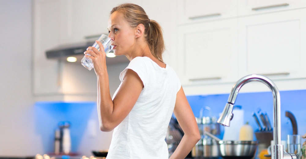 Frau trinkt Wasser aus einem Glas in der sauberen hellen Küche mit Küchengeräten und weißblauer Einrichtung.