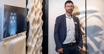 Mann steht in einem Raum zwischen strukturierten weißen Wänden und einem Flachbildfernseher der ein architektonisches Design zeigt. Der Mann trägt eine Brille ein Hemd und ein Sakko.