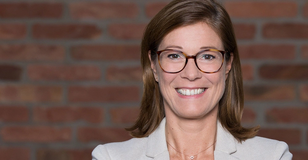 Frau trägt Brille und lächelt leicht vor einer Wand aus roten Backsteinen