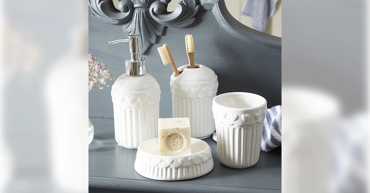Weiße keramische Badezimmerset bestehend aus einem Seifenspender zwei Zahnbürstenhaltern und einem Becher auf einem grauen Tisch mit Blumenvase links im Hintergrund. Seife liegt auf einer Seifenschale.