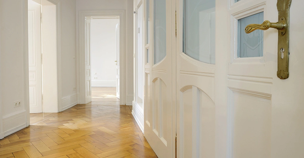 Tür geöffnet offener Flur holzgefertigter Fußboden weiße Wände und Türen heller Raum im Hintergrund Lichteinfall Steckdosen an der Wand.