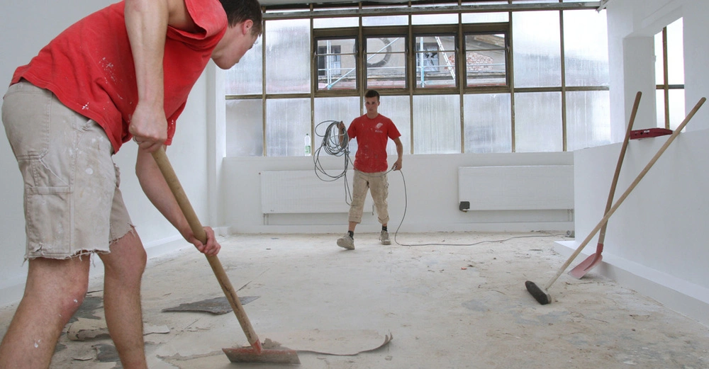 Zwei junge Männer, beide in einem roten T-Shirt und einer kurzen beigen Arbeitshose, bereiten den Estrich-Untergrund vor. Einer arbeitet mit einer Stoßscharre, der andere wickelt ein langes Kabel ab. Schaufel und Besen lehnen an der Wand. 