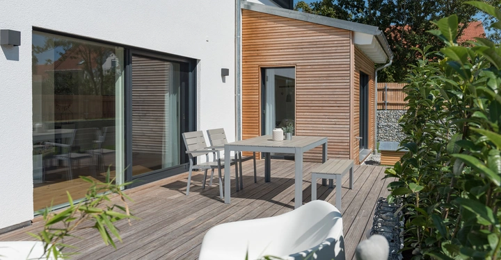 Terrasse mit Holzboden auf der ein Tisch mit Stühlen und einer Bank steht umgeben von Büschen und neben einem modernen weißen Haus mit großen Glastüren und einem Holzschuppen im Hintergrund