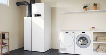 Waschmaschine und Trockner stehen nebeneinander auf einem gefliesten Boden. Rechts gibt es weiße Regale mit Handtüchern. Links steht ein hoher weißer Kessel, oben dekoriert mit Pflanzen; darüber ein Regal.