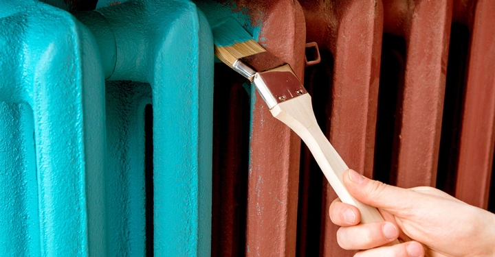 Frau, man sieht nur die rechte Hand, bemalt mit Heizkörperpinsel einen rostroten Heizkörper mit petrolfarbenem Lack