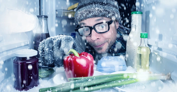 Mann trägt Winterkleidung und schaut in einen Kühlschrank gefüllt mit Gemüse und Gewürzen. Umgebung zeigt Schneeeffekt und frostige Bedingungen.